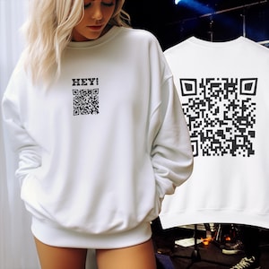 Puede incluir: Sudadera blanca con un código QR negro y el texto "HEY!" impreso en la parte delantera. La parte posterior de la sudadera tiene un código QR negro más grande.