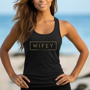 Puede incluir: Camiseta de tirantes negra con un gráfico dorado que dice "WIFEY EST. 07.08.25" en un cuadro.
