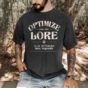 Op de afbeelding: Een zwart T-shirt met een vintage-stijl grafische afbeelding met de tekst "Optimize for the Lore" met daaronder de woorden "turn setbacks into legends". De afbeelding bevat ook de tekst "JAWDROP-147 2023" en "NATE & JONES".