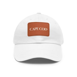 May include: White baseball cap with a brown rectangular patch. The patch features the words "CAPE COD" in white capital letters. The hat has a curved brim and a classic six-panel design.