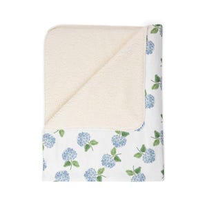 Puede incluir: Una manta blanca con un estampado floral de hortensias azules en un lado y un forro de forro polar sherpa color crema. La manta está doblada, mostrando las texturas y colores contrastantes.