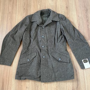 Puede incluir: Chaqueta de lana gris con cuello con solapas, cuatro botones y dos bolsillos delanteros. La chaqueta tiene mangas largas y una etiqueta en la parte inferior derecha. La chaqueta es de color neutro y parece vintage.