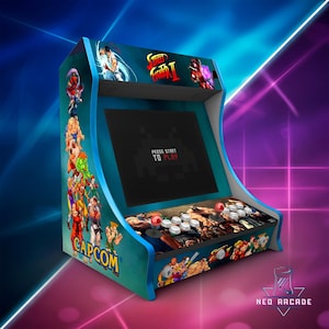 Peut inclure: Une borne d'arcade turquoise avec des graphismes de Street Fighter II et le logo Capcom. L'écran noir affiche "PRESS START TO PLAY" et un panneau de commande avec des joysticks et des boutons. Décorée d'illustrations de personnages.