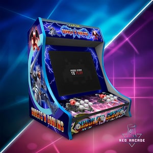 Puede incluir: Una máquina recreativa azul con el juego "Ghosts 'n Goblins" en la pantalla. El mueble tiene un diseño azul y blanco con gráficos de rayos y el texto "Press Start to Play". Cuenta con joysticks y botones.