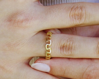 May include: A gold-colored ring with a repeating heart-shaped design. The ring is worn on a finger, showcasing its delicate and intricate pattern. The ring's design is a continuous loop of heart shapes.