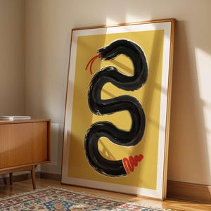 Può includere: Una stampa d'arte da parete incorniciata raffigurante un serpente nero audace, in stile pennellata, che si snoda verticalmente su uno sfondo giallo senape solido, con una lingua biforcuta rosso brillante in alto e un sonaglio rosso in fondo al corpo.