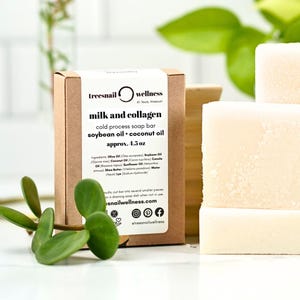 May include: A brown cardboard box with a white label displays the text "milk and collagen cold process soap bar." Beside the box are several stacked, rectangular, off-white soap bars. The label also lists ingredients and the weight, approximately 4.5 oz.