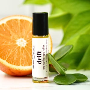 May include: Aromatherapy roller bottle with a black cap and white label that reads "Drift aromatherapy roller" with "cedarwood & chamomile - soothing sleep" text. An orange slice and green leaves are in the background.