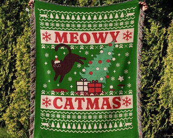 May include: Green woven blanket with a festive "Meowy Catmas" design. Features a black cat wearing a Santa hat, a Christmas tree, gifts, and snowflakes. The blanket has a fringed edge and a repeating pattern.