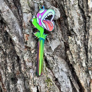 May include: A colorful pendant necklace featuring a stylized Piranha Plant design. The pendant has a rainbow-colored stem, a green and pink head with a red tongue, and white teeth. The necklace hangs against a textured tree bark background.
