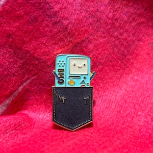 May include: A gold-colored pin of a blue and white video game character, BMO, with a smiling face, wearing a black pocket. The character is from the animated television series Adventure Time.