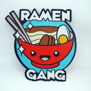 May include: A blue and white embroidered patch with a red bowl of ramen noodles. The bowl has a smiling face and the text "Ramen Gang" is written above and below the bowl.