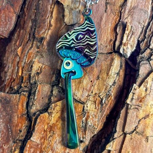 May include: A iridescent blue and black mushroom pendant necklace with a glittery top and a long, thin stem. The pendant is shaped like a cartoon mushroom with a single eye and a tongue sticking out.