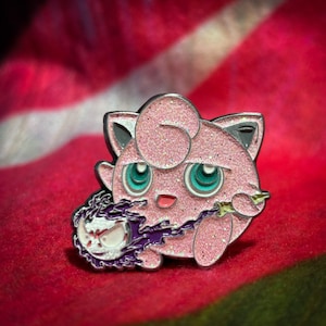 May include: A pink and silver glitter enamel pin of a Pokemon character,  Jigglypuff, holding a purple and white ghost-like object.