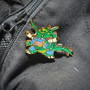 May include: A green and blue enamel pin featuring a cartoon Pokemon character with a jetpack. The character has a green body, blue arms and legs, and a yellow face with a pink tongue sticking out.