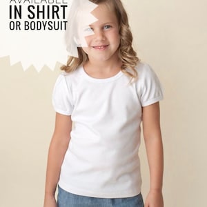 May include: A young girl wearing a white short-sleeved t-shirt and blue denim shorts. The text "AVAILABLE IN SHIRT OR BODYSUIT" is visible in the image.