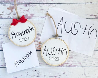 May include: Two embroidered hoop ornaments with names and the year 2023. One has the name "Hannah" with a red bow, the other says "Austin". Both have a twine loop for hanging.