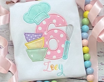 May include: A white onesie with a pink number six with white polka dots, a blue chef's hat, a set of pastel-colored mixing bowls, a whisk, and the name "Loey" embroidered on it.
