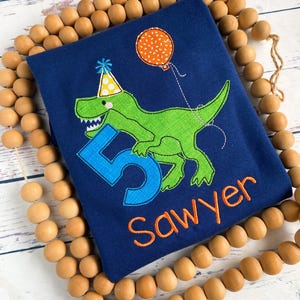 May include: Navy blue birthday shirt featuring a green dinosaur wearing a party hat and holding a balloon. The number 5 and the name "Sawyer" are embroidered in blue and orange, respectively. The shirt is surrounded by wooden beads.