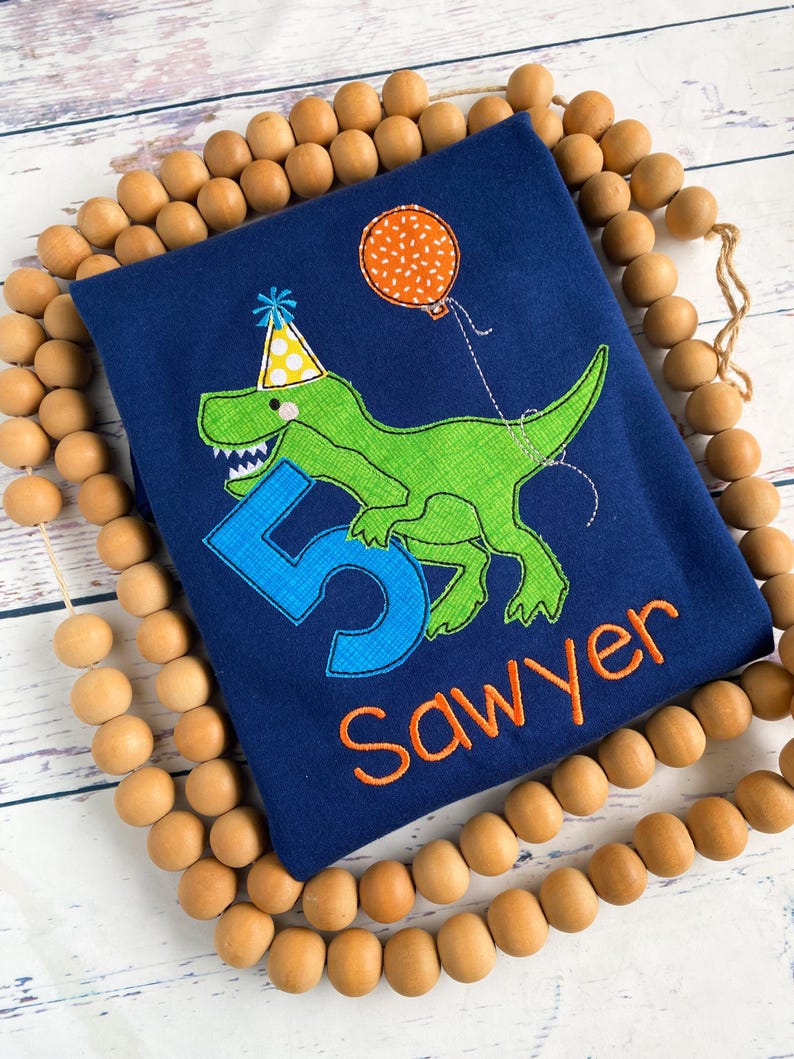 May include: Navy blue birthday shirt featuring a green dinosaur wearing a party hat and holding a balloon. The number 5 and the name "Sawyer" are embroidered in blue and orange, respectively. The shirt is surrounded by wooden beads.