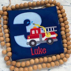 May include: Navy blue toddler shirt with a light blue number 3 applique. A red and gray fire truck with a yellow hose is embroidered on the shirt. The name "Luke" is embroidered in red below the fire truck.