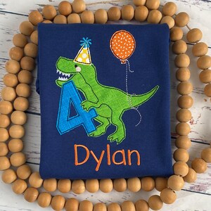 May include: Navy blue long-sleeve shirt featuring a green dinosaur wearing a party hat and holding a blue number 4. An orange balloon is attached to the dinosaur. The name "Dylan" is embroidered in orange.