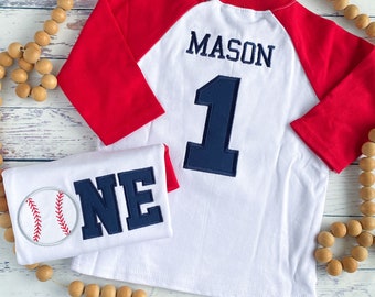 May include: A white baseball-themed baby outfit with red sleeves. The shirt has the name "MASON" and the number "1" embroidered on the back. The onesie has a baseball and the word "ONE" embroidered on the front.