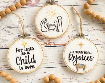 May include: Three embroidered Christmas ornaments with a white background and black embroidery. The first ornament says "For unto us a child is born". The second ornament depicts the nativity scene with a star above. The third ornament says "The weary world rejoices".
