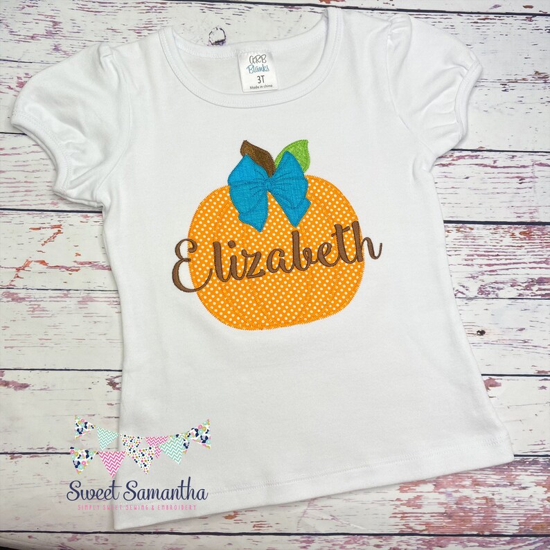 Puede incluir: Camiseta blanca de manga corta con una calabaza naranja con un lazo azul y el nombre "Elizabeth" bordado.