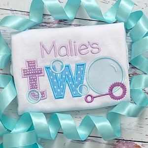 May include: A white onesie with the text "Mallie's Two" embroidered in purple, blue, and white. The number two is decorated with blue and white polka dots and surrounded by blue and white bubbles. A purple and white bubble wand is also embroidered on the onesie.