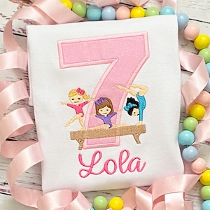 May include: A pink number seven with three girls in gymnastics poses on a brown balance beam. The name "Lola" is embroidered below the number.