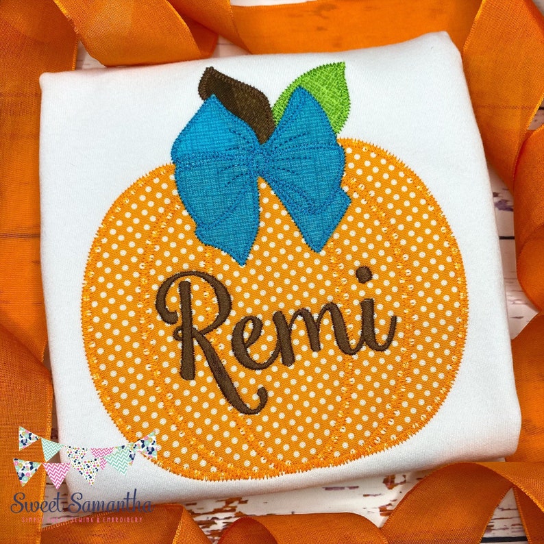 Puede incluir: Una camiseta blanca con una calabaza naranja con lunares blancos y un lazo azul. El nombre "Remi" est&aacute; bordado en la calabaza.