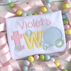 May include: A white onesie with the text "Violet's Two" embroidered in pink, yellow, and blue. The number two is decorated with bubble designs.