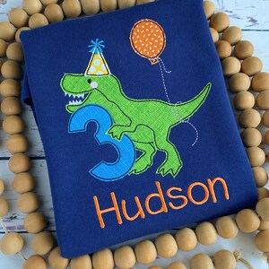 May include: Navy blue toddler shirt with an embroidered green dinosaur wearing a party hat and holding a balloon. The number 3 and the name "Hudson" are also embroidered in blue and orange.