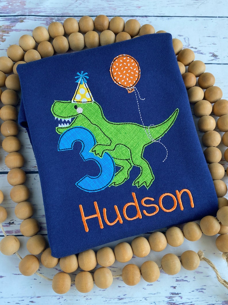 May include: Navy blue toddler shirt with an embroidered green dinosaur wearing a party hat and holding a balloon. The number 3 and the name "Hudson" are also embroidered in blue and orange.