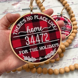 May include: A red and black buffalo plaid fabric hoop ornament with white embroidery that reads "There's no place like home 34472 for the holidays".