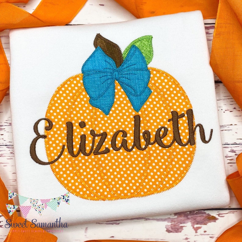 Puede incluir: Una camisa blanca con una calabaza naranja con lunares blancos y un lazo azul en la parte superior. El nombre "Elizabeth" est&aacute; bordado en cursiva marr&oacute;n sobre la calabaza.