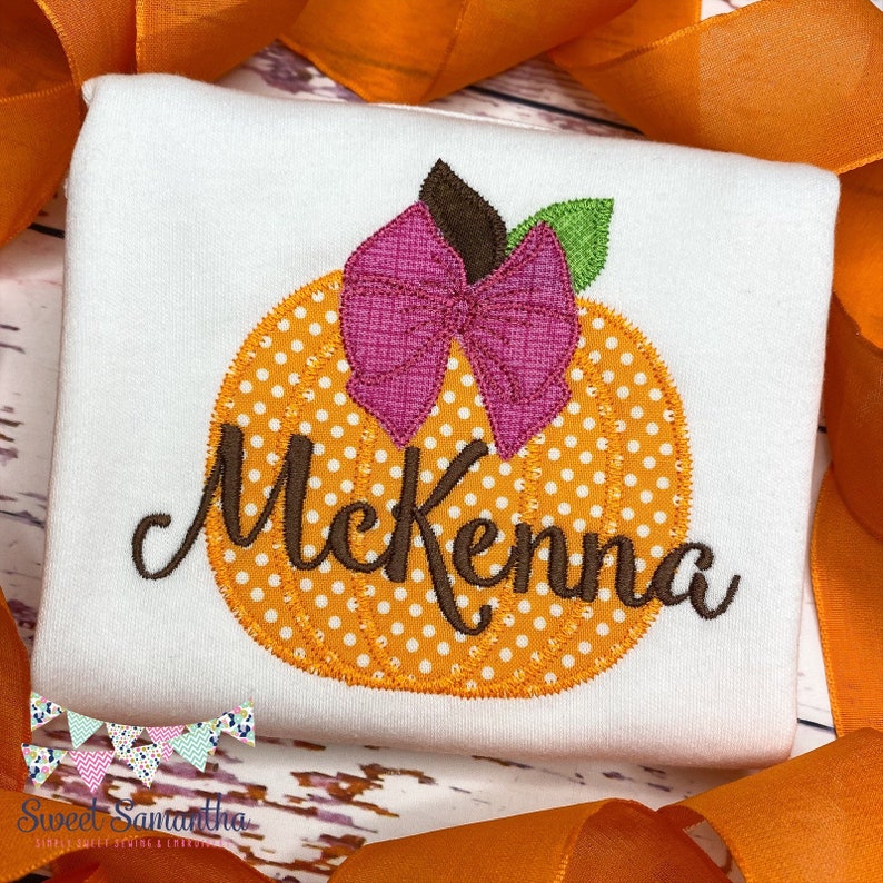 Puede incluir: Una camiseta blanca con una calabaza naranja con lunares blancos y un lazo rosa en la parte superior. El nombre McKenna est&aacute; bordado en marr&oacute;n debajo de la calabaza.