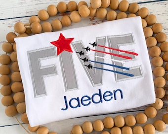 May include: White t-shirt with the word "FIVE" in gray, featuring a red star and jets with red, white, and blue trails. The name "Jaeden" is embroidered in blue below the number. The shirt is surrounded by wooden beads.