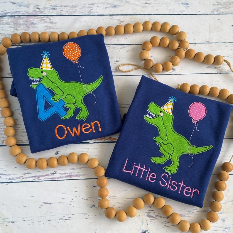 May include: Two navy blue t-shirts with dinosaur designs. One shirt has the name "Owen" and the number "4" embroidered in blue and orange. The other shirt says "Little Sister" in pink. Both shirts feature a green dinosaur wearing a party hat and holding a balloon.