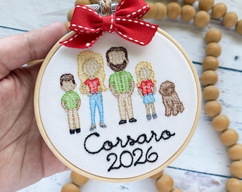 May include: A circular embroidery hoop featuring a stitched family portrait of four people and a brown dog on white fabric. A red ribbon bow with white stitching sits at the top. The text "Corsaro 2026" is embroidered in black script below the figures.