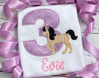 Puede incluir: Una camiseta blanca con un número 3 de purpurina morado y un caballo marrón con una crin y cola negras. El nombre "Evie" está bordado en rosa debajo del caballo.