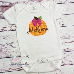 Puede incluir: Mono blanco para beb&eacute; con una calabaza naranja con lunares blancos, un lazo rosa y un tallo verde. El nombre McKenna est&aacute; bordado en marr&oacute;n en la calabaza.
