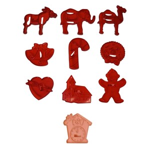 May include: A collection of red plastic cookie cutters in various shapes, including an elephant, camel, donkey, bells, candy cane, wreath, heart, church, gingerbread man, and a cuckoo clock.