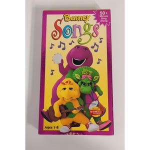 May include: A VHS tape titled "Barney Songs" featuring Barney the dinosaur and friends. The cover is yellow with musical notes and the characters are purple, green, and yellow. The tape is labeled "50+ Minute Sing Along" and is for ages 1-8.