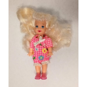 May include: A small doll with blonde hair and blue eyes. The doll is wearing a pink and white checkered outfit with floral accents and a white bow. The doll is wearing pink shoes.