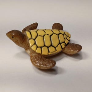 May include: A small, detailed turtle figurine. The turtle has a yellow shell with black outlines, brown head, legs, and tail. The figurine is made of a glossy material and is set against a white background.