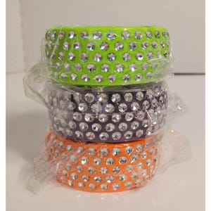 May include: Stack of three colorful bracelets: lime green, purple, and orange. Each bracelet is adorned with numerous clear, sparkling rhinestones. The bracelets are stacked on top of each other and wrapped in clear plastic.
