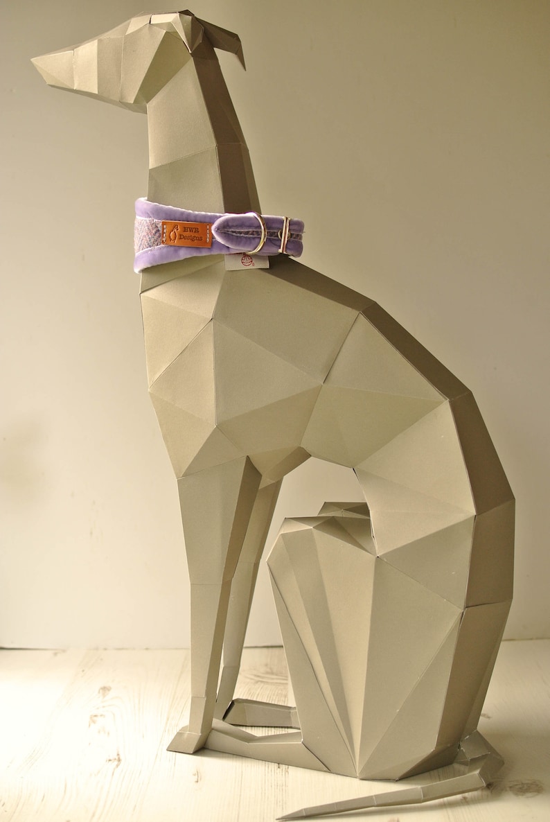 May include: A light gray paper model of a dog sitting with its tail curled under its body. The dog is wearing a lavender velvet collar with a brown leather tag that says "Elvie Designs".