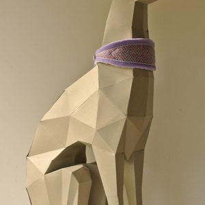 May include: A beige papercraft sculpture of a greyhound dog sitting with its legs crossed. The dog is wearing a purple and pink patterned collar.